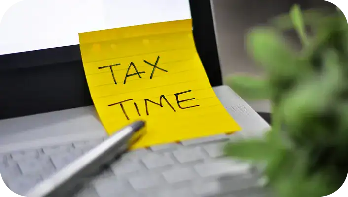 Yellow sticky note reading “Tax Time” on a laptop keyboard with pen and blurred plant in foreground.